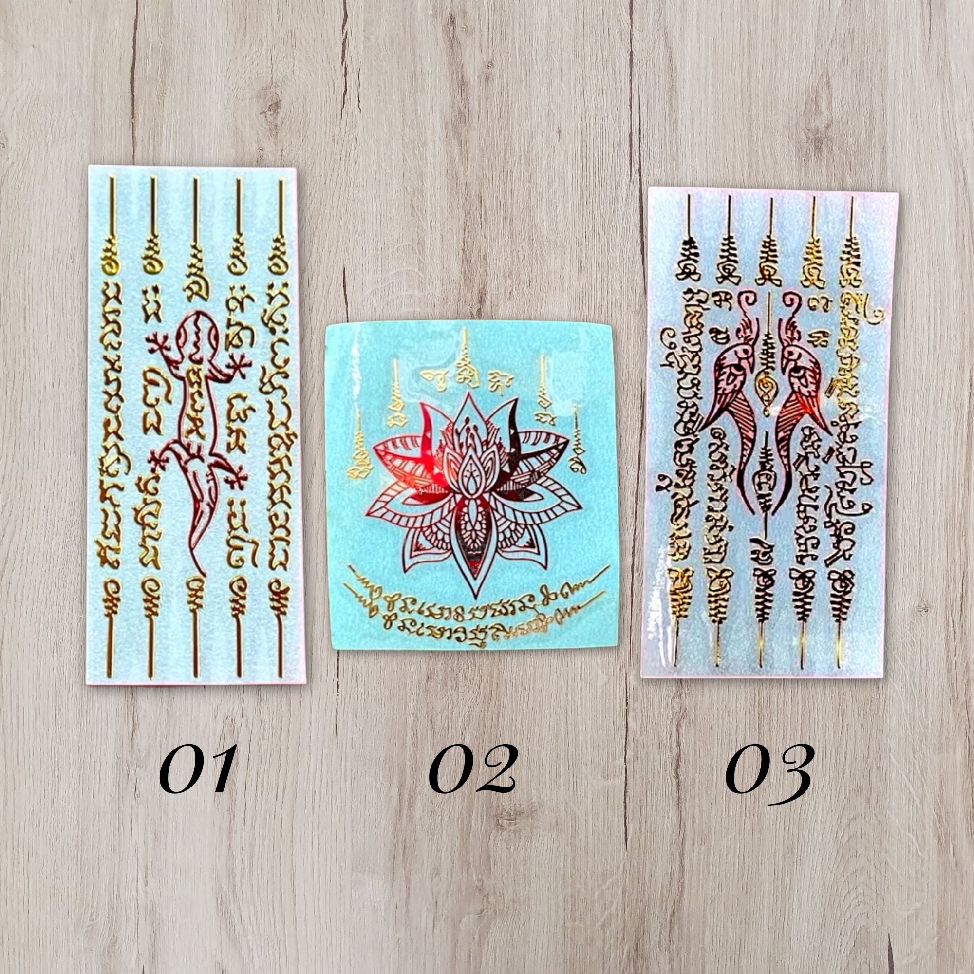 Three temporary tattoo sticker sheets displayed on a light wood background, labeled 01, 02, and 03. The designs feature gold-toned Thai script and traditional motifs including a red and gold lotus flower and a red and gold lizard on light blue and white backing papers.